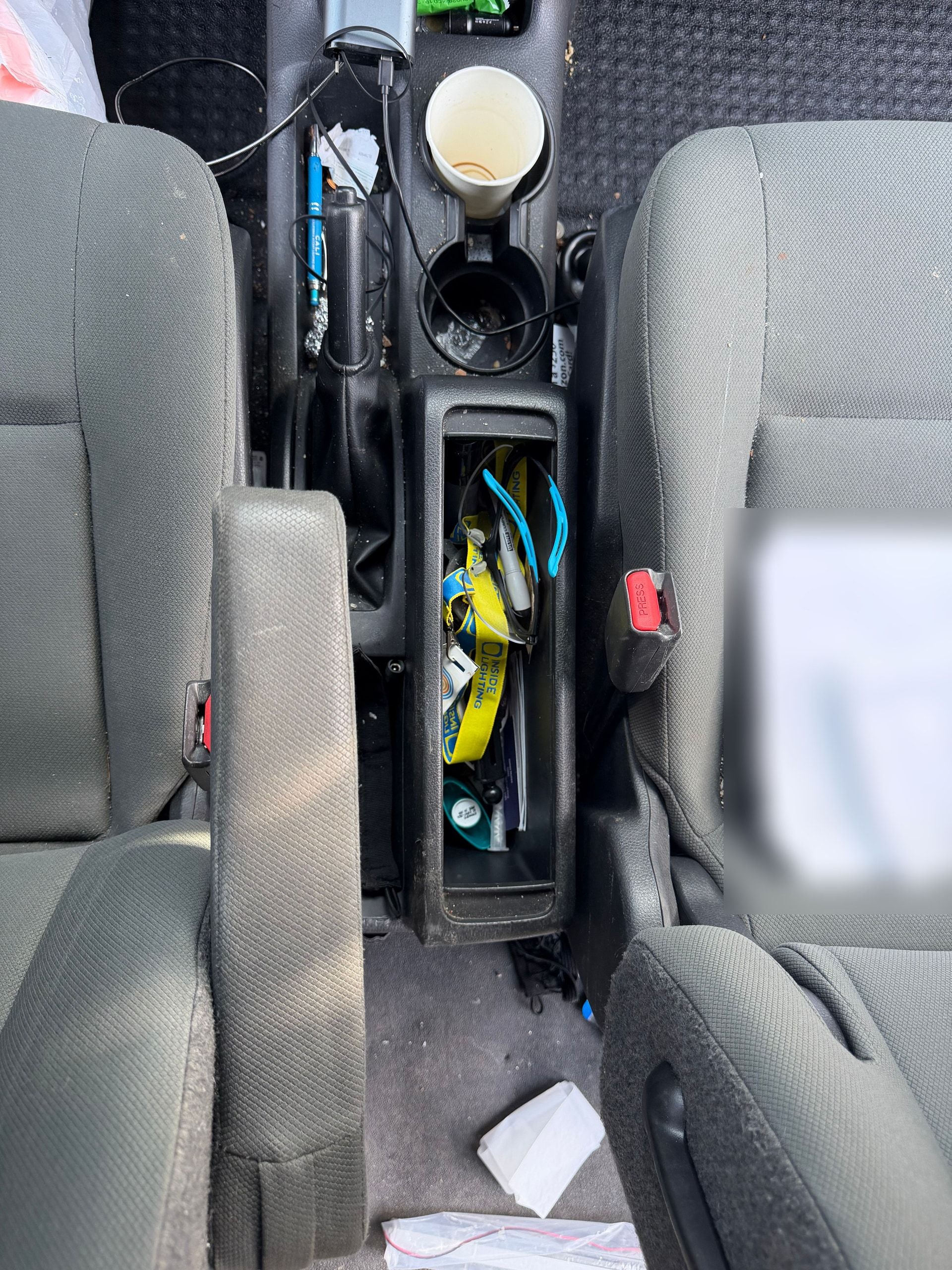 Nissan NV200 front seats and center console before detailing, with trash, loose cables, a used cup and debris on the floor.